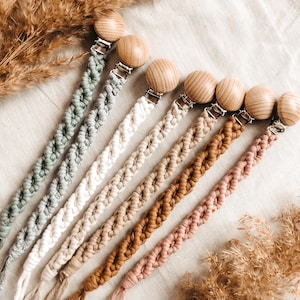 Könnte beinhalten: Ein Set aus acht Schnullerketten mit Holzperlen in verschiedenen Naturfarben, darunter Beige, Braun und Rosa. Die Ketten sind aus geflochtenem Baumwollseil gefertigt und haben einen silbernen Metallclip.