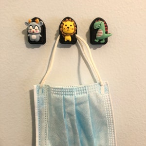 May include: Three colorful animal-shaped hooks with a blue, yellow, and green design are mounted on a white wall. A light blue face mask is hanging from the yellow lion hook.