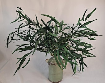 May include: A green ceramic vase with two handles holds a lush arrangement of green eucalyptus leaves and small white flowers. The vase has a distressed, aged appearance with brown accents, creating a rustic aesthetic. The leaves are long and slender, creating a full, natural display.