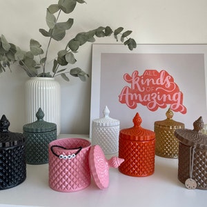 May include: A selection of diamond-patterned, lidded jars in shades of black, green, pink, white, orange, gold, and brown. The jars feature decorative finials. One pink jar is open, displaying the word "MAMA" on a beaded strand. A framed print reads "ALL KINDS OF AMAZING."