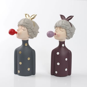 Puede incluir: Dos figuritas decorativas de personas soplando burbujas. La figurita de la izquierda lleva un vestido de lunares grises y tiene una burbuja roja. La figurita de la derecha lleva un vestido de lunares marrones y tiene una burbuja morada.
