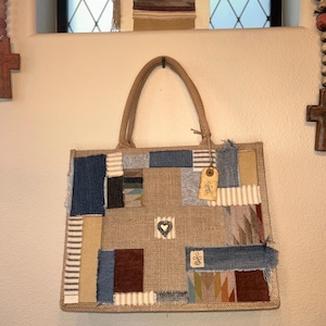 Könnte beinhalten: Eine Patchwork-Tragetasche mit Jutebasis und Denim-, Streifen- und gemusterten Stoffflicken. Die Tasche hat eine Herz- und Blumenverzierung und ein Etikett. Die Griffe bestehen aus gewebtem Material.