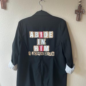 Può includere: Blazer nero con maniche arrotolate, con la scritta "ABIDE IN HIM 1 JOHN 2:28" sul retro. Il testo è in stile patchwork con accenti bianchi, rosa e beige. Il blazer è appeso a una gruccia di metallo.