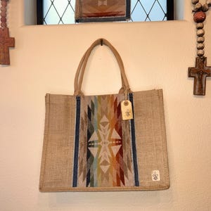 May include: A beige jute tote bag with a colorful geometric design on the front. The bag has sturdy handles and a small tag. The design features a mix of colors including blue, green, orange, and red. A small logo is visible on the bottom right corner.