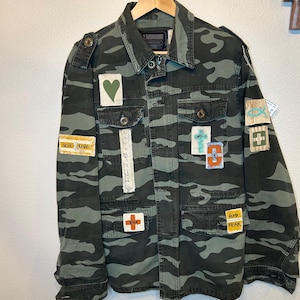 Puede incluir: Chaqueta con estampado de camuflaje con múltiples parches. Los parches incluyen un corazón verde, una cruz y textos como "TRUE LOVE", "GOD FEAR" y "KINA". La chaqueta está colgada en una percha de madera.