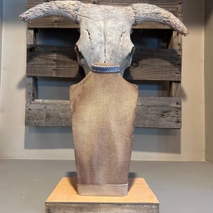 May include: A brown burlap jewelry display bust with a blue and white patterned necklace. The bust is on a wooden base, with a weathered wooden pallet and a cow skull in the background.