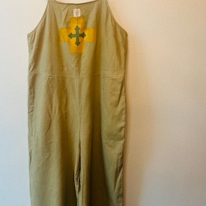 May include: Khaki-colored jumpsuit with a yellow and green cross design on the chest. The jumpsuit has wide legs and adjustable straps. The garment is hanging on a wooden hanger against a white wall.