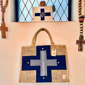 May include: A jute tote bag with a navy blue cross design, accented with silver studs. The bag has tan handles and a small tag. A matching decorative pillow with a cross design sits above the bag. Wooden rosary beads and crosses are on either side.