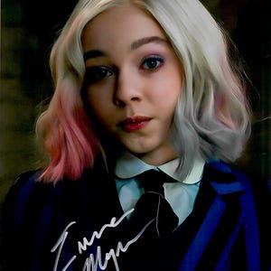 May include: A portrait of a person with light skin, light hair with pink highlights, and blue eyes. They are wearing a black blazer with blue stripes and a white collared shirt. The image includes a signature.