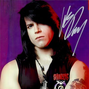 May include: A person wearing a black sleeveless shirt with a graphic on the front and a cross pendant. The person has long black hair and a tattoo on the arm. The background is purple.
