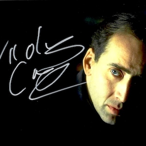 May include: A headshot of actor Nicolas Cage with his signature in white ink on a black background. The signature is in cursive and reads "Nicolas Cage". The actor is looking towards the viewer with a serious expression.