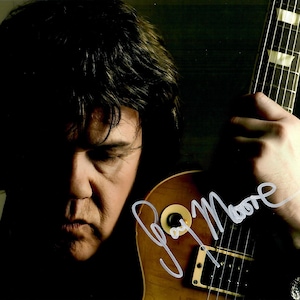 以下が含まれることがあります： ギターを持つ男性の写真で、白い「Gary Moore」のサイン入り。ギターは明るい茶色で、指板は黒です。男性は黒髪で、下を見ています。