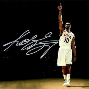 May include: A signed photograph of a basketball player in a USA jersey, number 10, raising his arm. The player is wearing white shorts and shoes. The background is dark with a signature in the upper left corner.