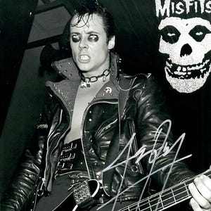 May include: Black and white photograph of a person in a leather jacket, playing a black bass guitar. The person is wearing heavy eye makeup and a spiked collar. A Misfits logo is visible in the background.
