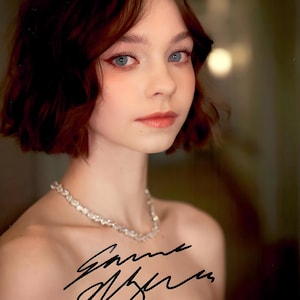 May include: A portrait of a person with fair skin, blue eyes, and short, auburn hair. They are wearing a sparkling necklace and have a signature in black ink on their chest. The background is blurred, with warm tones.