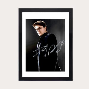 Puede incluir: Fotografía en blanco y negro enmarcada de una persona con gafas y un traje oscuro, con una firma blanca. La fotografía está sobre un fondo oscuro y enmarcada en un marco negro.