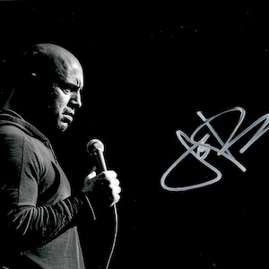 Peut inclure: Photographie en noir et blanc d'un homme tenant un microphone, avec une signature dans le coin supérieur droit. L'homme porte une chemise sombre à manches longues et regarde sur le côté. Le fond est noir.
