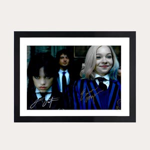 May include: Framed print showing three people in formal wear. Two young women are the focus, one with dark hair and the other with blonde hair, both wearing dark blazers, white shirts and ties. The print is set against a neutral background.