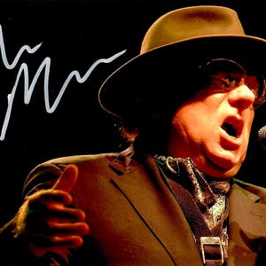 Puede incluir: Un hombre con un sombrero fedora marrón y una chaqueta de traje marrón canta en un micrófono. Lleva una bufanda con estampado blanco y negro alrededor del cuello. La imagen está firmada con el nombre "Jim".