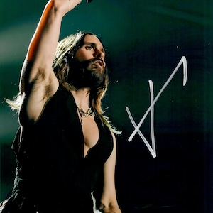 May include: A person with long hair and a beard, wearing a black vest and a necklace, raises their arm. A tattoo is visible on the arm. The background is dark with a white signature.