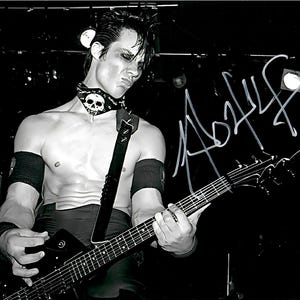May include: Black and white photograph of a person playing an electric guitar on stage. The person is wearing a skull-adorned choker and armbands. The image is signed in white ink.