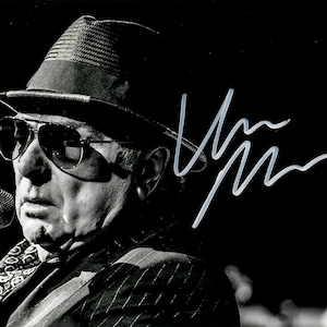 Puede incluir: Fotografía en blanco y negro de una persona con sombrero fedora, gafas de sol de aviador y una bufanda estampada. La persona sostiene un micrófono y hay una firma en la esquina superior derecha.