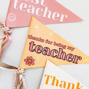May include: Three colorful pennant flags with text for teachers. The pink flag says "Best teacher" with a smiley face. The orange flag says "thanks for being my teacher" with a flower. The white flag says "Thank You!"