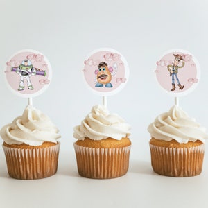 K&ouml;nnte beinhalten: Drei Cupcakes mit wei&szlig;em Zuckerguss, jeweils gekr&ouml;nt mit einer runden Dekoration mit einer anderen Figur aus dem Animationsfilm Toy Story: Buzz Lightyear, Mr. Potato Head und Woody. Die Dekorationen sind vor einem rosa Hintergrund mit Wolken.