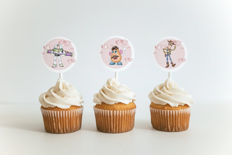 K&ouml;nnte beinhalten: Drei Cupcakes mit wei&szlig;em Zuckerguss, jeweils gekr&ouml;nt mit einer runden Dekoration mit einer anderen Figur aus dem Animationsfilm Toy Story: Buzz Lightyear, Mr. Potato Head und Woody. Die Dekorationen sind vor einem rosa Hintergrund mit Wolken.