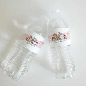 K&ouml;nnte beinhalten: Zwei transparente Plastikwasserflaschen mit Etiketten mit Zeichentrickfiguren und dem Text "Happy Birthday Amelia". Die Etiketten haben einen rosa Hintergrund mit Cartoon-Illustrationen. Die Flaschen sind mit klarer Fl&uuml;ssigkeit gef&uuml;llt.