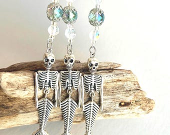 May include: Three silver-toned skeleton mermaid charms hanging from vertical strands of clear, iridescent, and faceted glass beads. Each charm features a detailed skull, a ribcage, and a fish tail, suspended by metal jump rings and chains.
