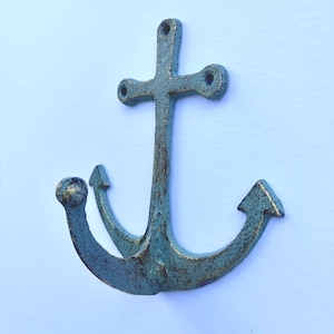 May include: A wall-mounted anchor hook featuring a textured, weathered teal and gold finish. The metal object has a central vertical shank with two mounting holes at the top and two curved arms extending outward, each ending in a pointed fluke.