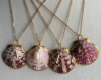Puede incluir: Cuatro collares de cadena dorada, cada uno con un colgante de concha natural con patrones morados y blancos. Tres collares incluyen un dije de letra dorada, J, L y C, mientras que un collar presenta un pequeño dije de perla blanca.