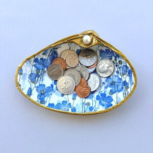May include: A decorative clam shell dish with a gold-painted rim and a white pearl accent. The interior features a blue and white floral pattern and contains a collection of assorted United States coins including quarters, dimes, and pennies.