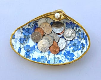 May include: A decorative clam shell dish with a gold-painted rim and a white pearl accent. The interior features a blue and white floral pattern and contains a collection of assorted United States coins including quarters, dimes, and pennies.