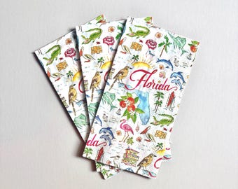 May include: Three white paper napkins featuring a colorful illustrated map of Florida. The design includes drawings of alligators, pelicans, flamingos, dolphins, palm trees, orange blossoms, and lifeguard towers, with the word Florida in red script.