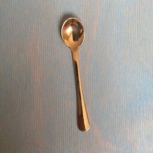 May include: A polished rose gold-toned metal spoon with a smooth, reflective surface and a classic oval bowl. The spoon features a slender, tapered handle that widens slightly at the base, showcasing a clean, minimalist design with no patterns.
