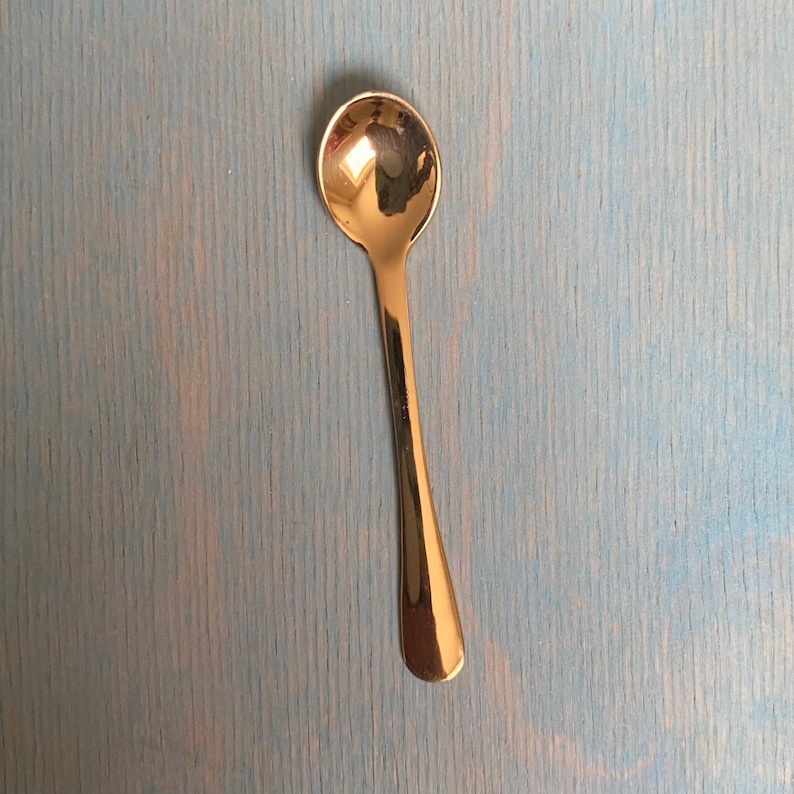 May include: A polished rose gold-toned metal spoon with a smooth, reflective surface and a classic oval bowl. The spoon features a slender, tapered handle that widens slightly at the base, showcasing a clean, minimalist design with no patterns.