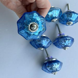 May include: A collection of six blue glass cabinet knobs with a crackled texture and a faceted, star-like shape. Each knob features a central silver-toned metal hardware component and a threaded metal bolt extending from the back for installation.
