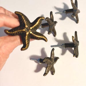 May include: A set of five starfish-shaped cabinet knobs featuring a dark bronze finish with gold-toned highlights on the textured surface. Each knob includes a threaded metal bolt and a small nut for installation on furniture or cabinetry doors.