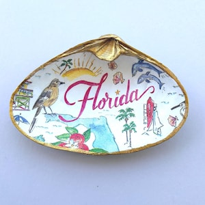 May include: A decorative seashell dish with a gold-painted rim, featuring a hand-painted illustration of Florida. The design includes a bird, a sun, palm trees, dolphins, a lifeguard tower, and the word Florida written in a pink cursive script.