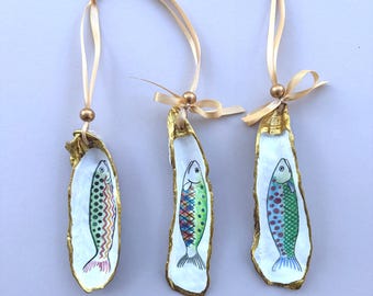 May include: Three oyster shell ornaments featuring hand-painted fish designs in vibrant colors. Each shell has a gold-painted edge and is suspended by a light beige ribbon with a small gold bead accent at the top of the shell.