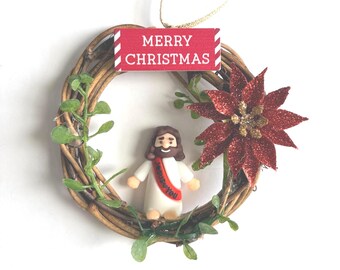 May include: A circular twig wreath ornament featuring a small figurine of a person with brown hair and a white robe with a red sash that says Jesus Loves You. A red sign reads MERRY CHRISTMAS, and a red glittery poinsettia flower is attached.