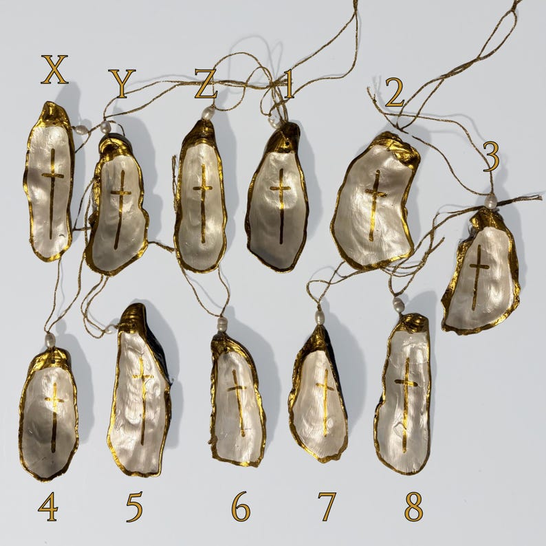 Puede incluir: Una colecci&oacute;n de doce adornos de concha de ostra, cada uno con una cruz pintada en oro en el interior, bordes dorados y una peque&ntilde;a cuenta blanca unida a un fino cord&oacute;n dorado. Las conchas est&aacute;n etiquetadas con las letras X, Y, Z, 1, 2, 3, 4, 5, 6, 7, 8.