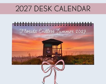 May include: A 2027 desk calendar with a black spiral binding. The cover features a sunset beach scene with a lifeguard tower and the text Florida Endless Summer 2027. A pink line-drawn bow is centered at the bottom of the calendar cover.