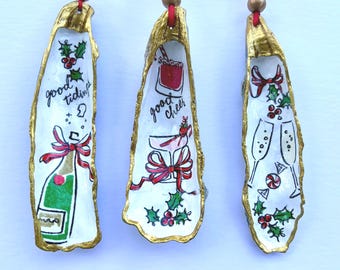 May include: Three oyster shell ornaments with gold-painted edges, each hanging from a red ribbon with a gold bead. The shells feature hand-painted holiday illustrations including a champagne bottle, cocktails, holly, and the text good tidings and good cheer.