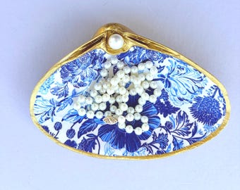 May include: A clam shell trinket dish featuring a blue and white floral pattern on the interior, with a metallic gold painted rim and a single white pearl accent at the top. A delicate white pearl necklace rests inside the shell.