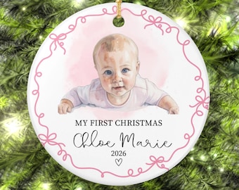 May include: A round white ceramic ornament featuring a watercolor illustration of a baby. The text reads MY FIRST CHRISTMAS, Chloe Marie, and 2026 with a small heart. A pink line drawing of a ribbon border frames the design.
