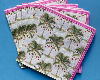 May include: A stack of white paper napkins with a tropical Christmas theme. Each napkin features palm trees decorated with ornaments. The napkins have a pink border with a striped design. The napkins are on a blue surface.