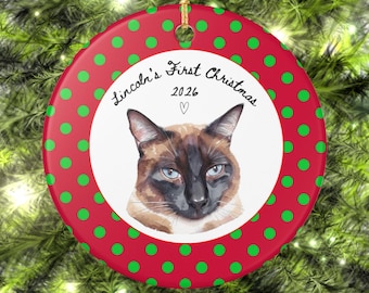 May include: A round ceramic ornament with a red background featuring green polka dots. The center white circle displays a portrait of a Siamese cat with blue eyes, the text "Lincoln's First Christmas 2026", and a small heart symbol.